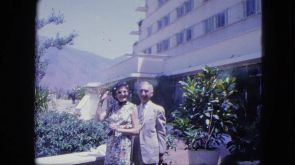 The woman raises a hand in a small wave as the couple smiles on the planted terrace, with mountains and hotel architecture behind them.