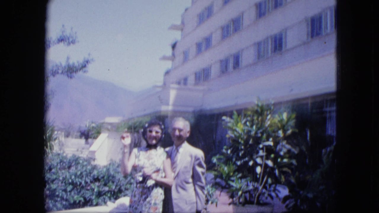 Woman waves beside a suited man outside a hotel with greenery and mountains.