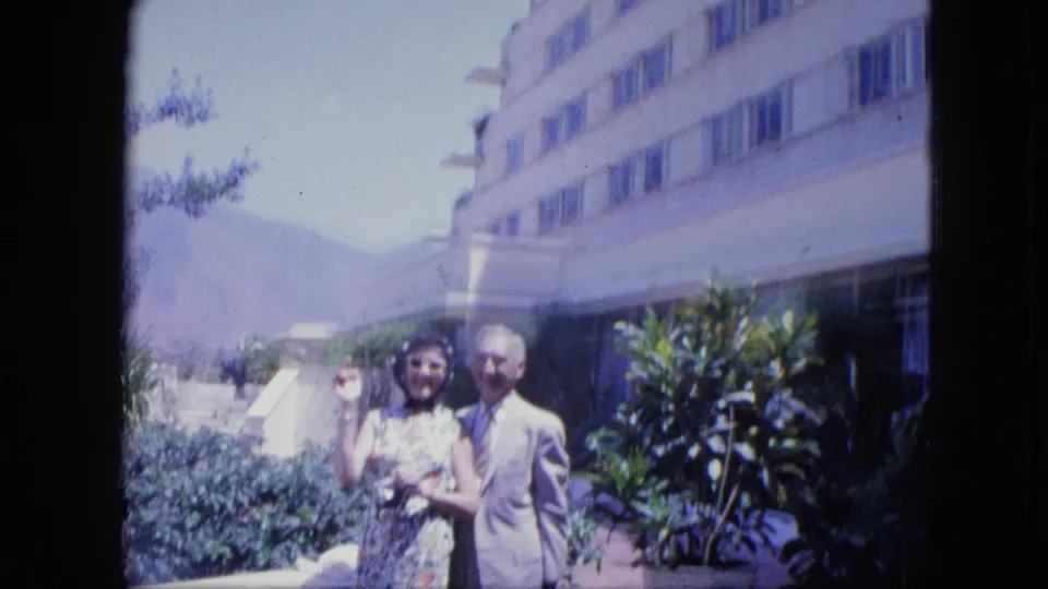 1964 Venezuela travel frame showing a woman waving next to a suited man on a hotel terrace with tropical plants and mountains visible.