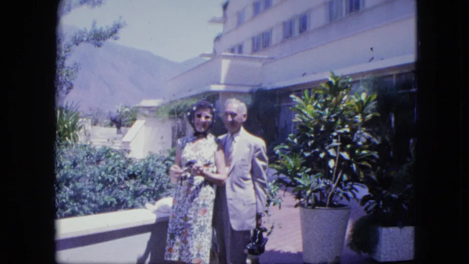 1964 Venezuela 8mm still of a couple standing arm in arm near a hotel terrace railing, framed by shrubs, potted plants, and mountains.