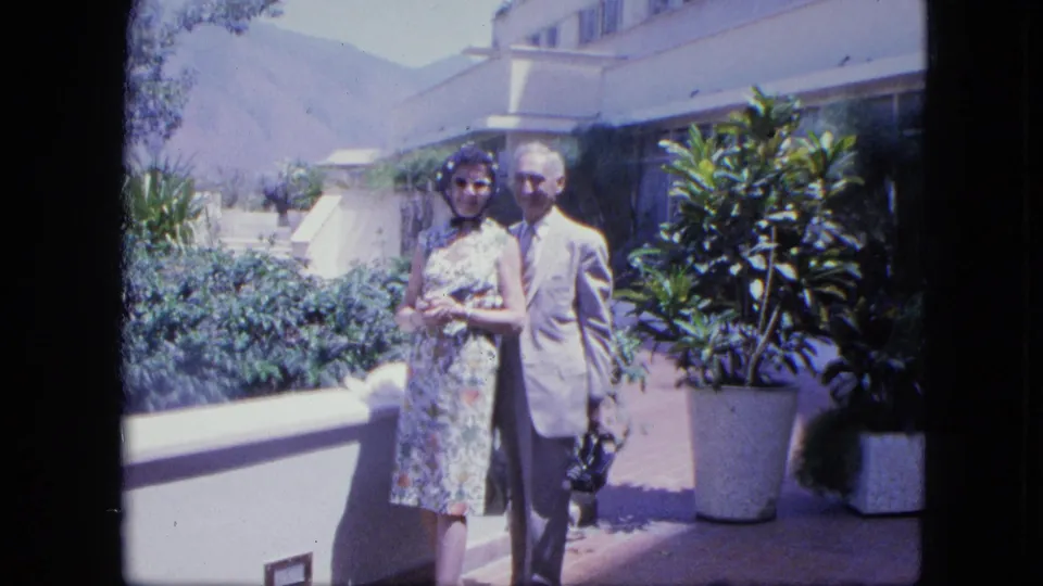 1964 Venezuela home-movie frame of a couple posed on a bright hotel terrace beside large potted plants with mountains beyond.