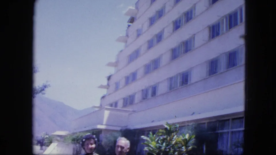 1964 Venezuela travel frame with the camera tilted toward a modern hotel facade while the couple remains partly visible below.