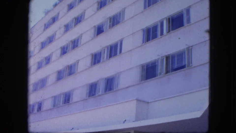 1960s 8mm frame of a white modern high-rise facade viewed from below, with dark film borders and soft blue color shift.