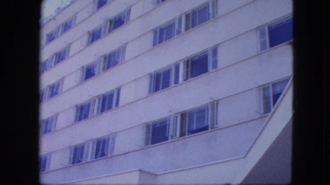 White modern building facade fills the frame, with repeated windows and slight handheld tilt in archival 8mm footage.