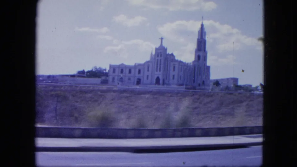 Distant cathedral-like landmark with a tall spire and cross, filmed from a moving vehicle in a blue-shifted 8mm travel shot.