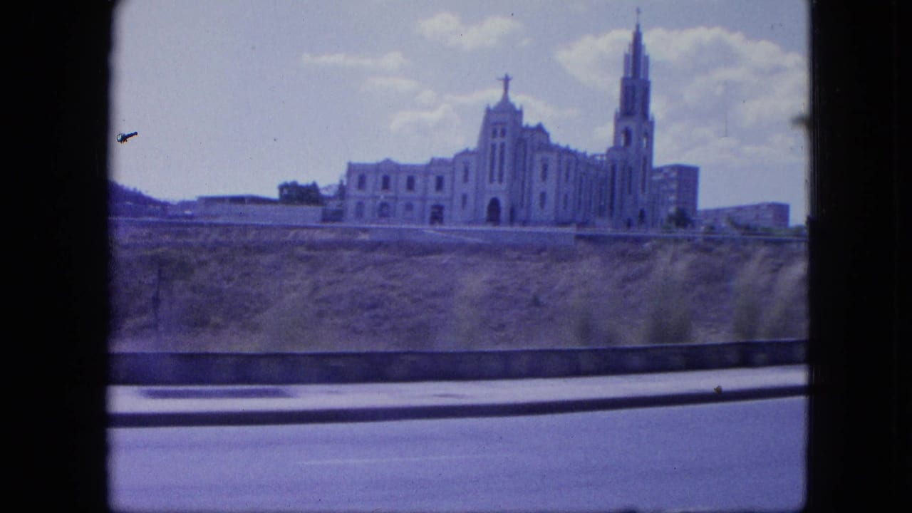 1964 Venezuela home-movie frame showing a large church-like building with twin spires and a cross, filmed from a moving car.