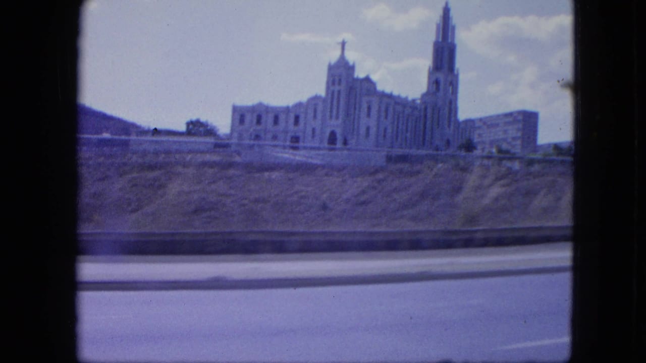1964 Venezuela travel frame with a church-like landmark and roadside barrier, softened by vehicle movement and 8mm color shift.