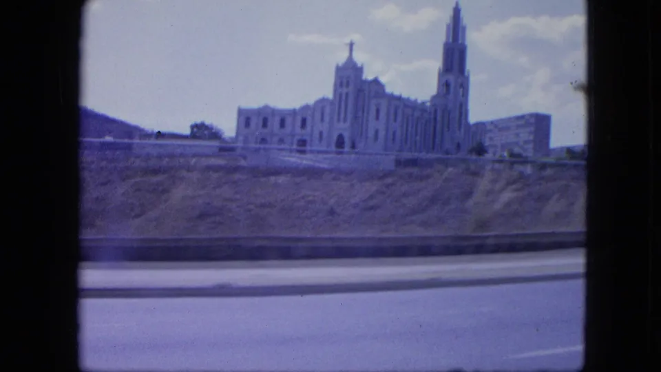 1964 Venezuela travel frame with a church-like landmark and roadside barrier, softened by vehicle movement and 8mm color shift.