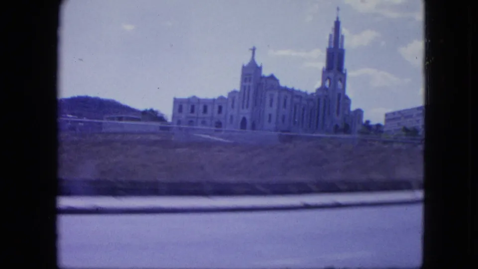 1964 Venezuela frame showing a cross-topped church-like facade and tall spire beyond a roadside embankment.