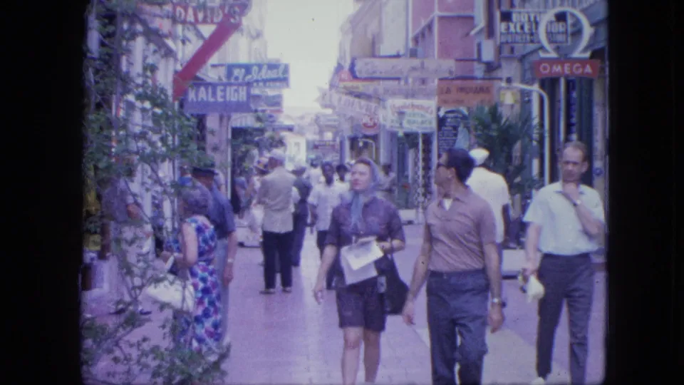 1964 Venezuela street scene with pedestrians, shopfronts, visible KALEIGH and OMEGA signs, and home-movie color grain.