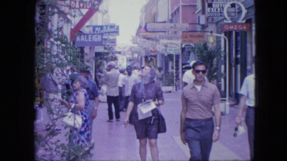 1964 8mm home-movie frame of pedestrians on a Venezuelan shopping street with Omega and Raleigh signs visible above the walkway.