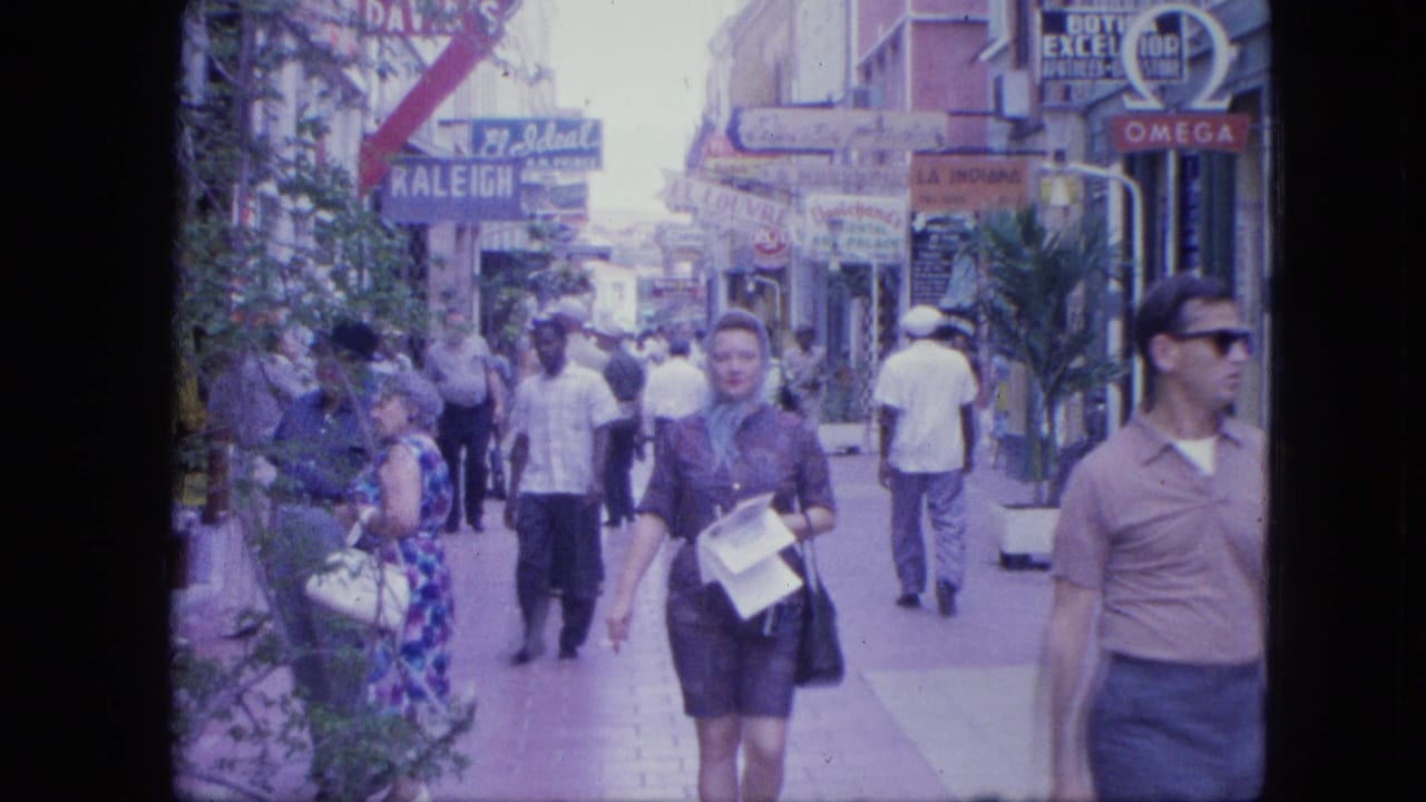 A woman holding papers walks toward the camera as pedestrians move past shops.