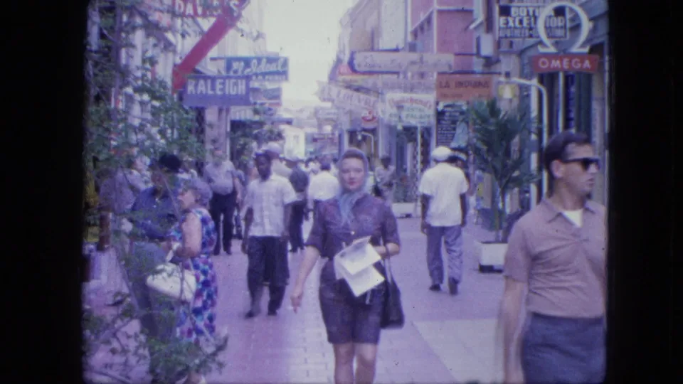 A woman with papers approaches the camera on a busy 1960s Venezuelan pedestrian street with storefront signs and passersby around her.