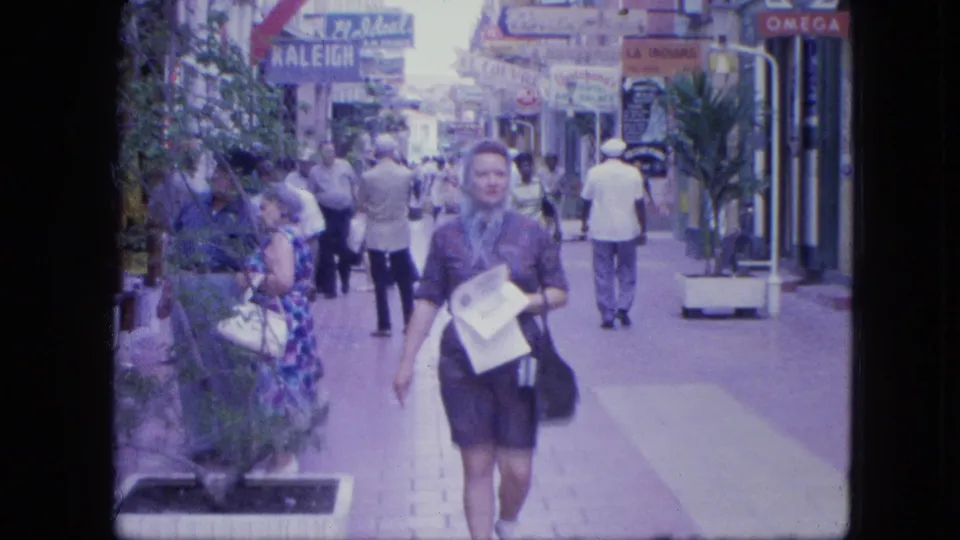 1964 home-movie frame showing a woman in a headscarf carrying papers on a Venezuelan shop-lined pedestrian walkway.