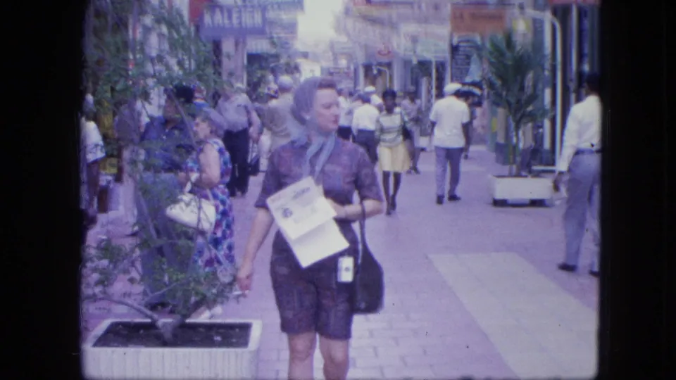 A woman with papers passes close to the camera on a 1960s Venezuelan pedestrian shopping street with visible storefront signage.