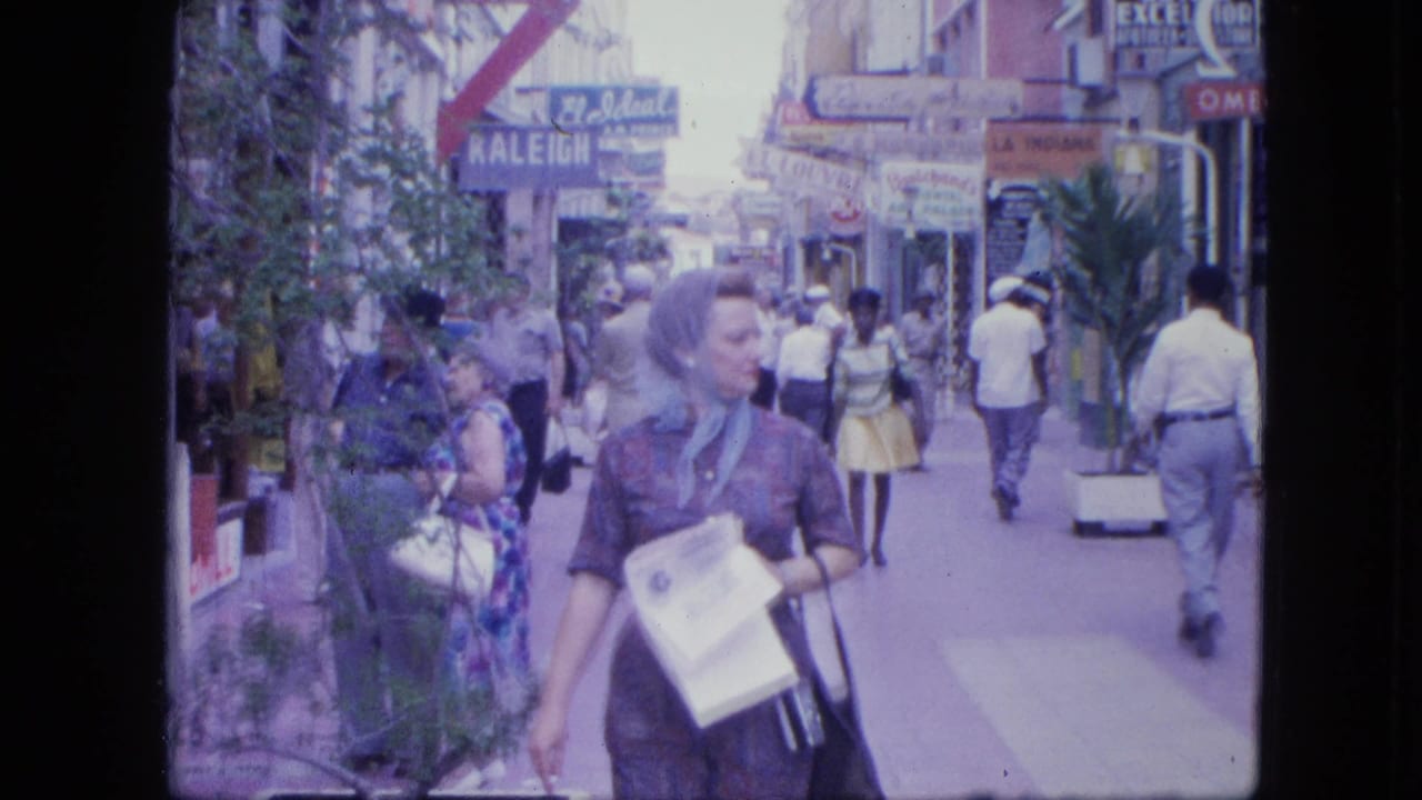 A woman carrying papers and a purse walks toward the camera on a busy 1960s shopping street with visible storefront signage.
