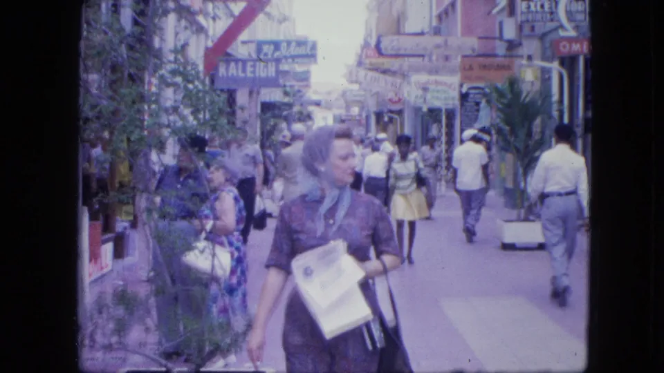 A woman carrying papers and a purse walks toward the camera on a busy 1960s shopping street with visible storefront signage.