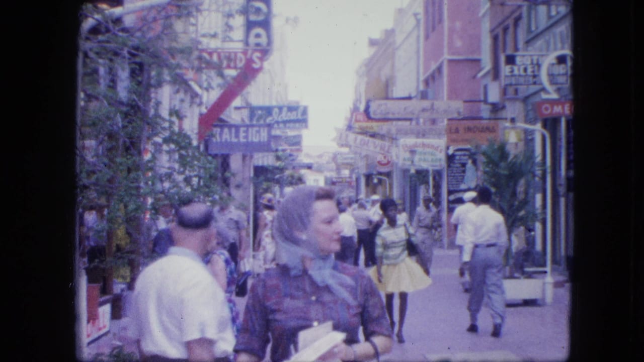 The woman in a patterned dress and foulard turns her head as pedestrians move behind her on a 1960s commercial street.