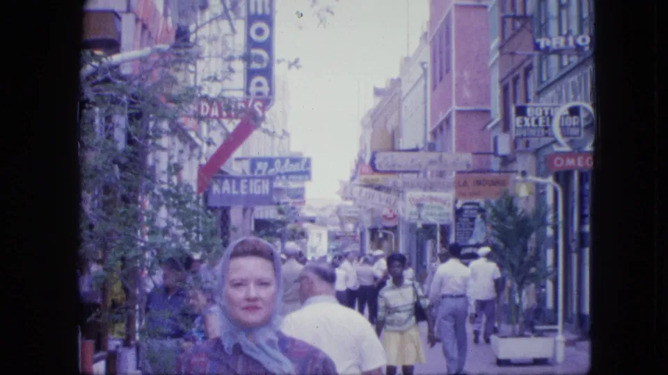 A woman with a light headscarf faces the camera near storefronts while people walk along a busy 1960s shopping street.