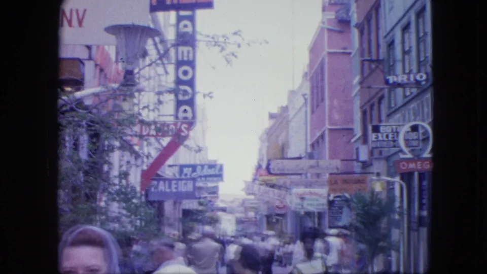 The camera holds on the commercial street as pedestrians, shop signs, and the partly cropped woman remain visible in the 8mm frame.