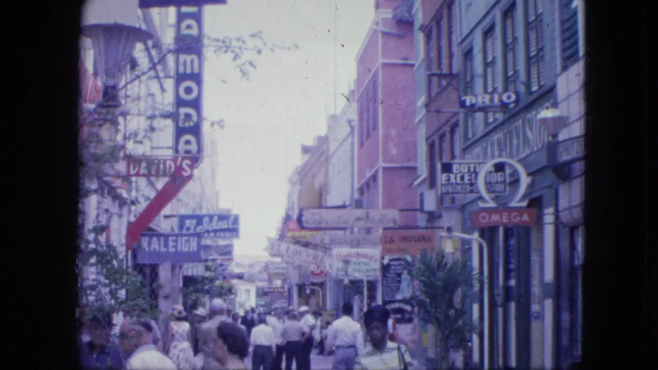 1964 Venezuela home-movie frame of pedestrians on a busy commercial sidewalk lined with storefronts and signs.