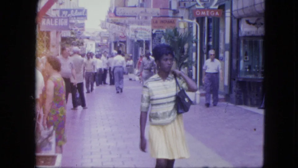 A woman passes close to the camera on a 1964 Venezuela shopping street with adult pedestrians and storefront signage behind her.
