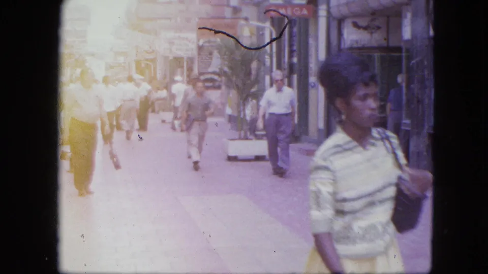 End frame of the 1964 Venezuela street clip, with bright exposure flare, pedestrians, storefronts, and visible commercial signs.