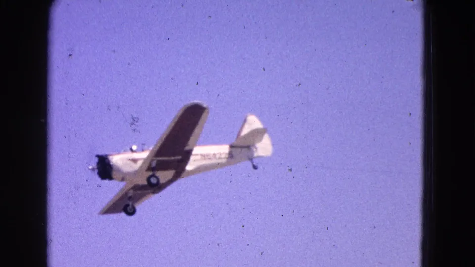 1964 8mm home-movie frame of a small single-engine airplane in flight against a pale sky, with landing gear visible.