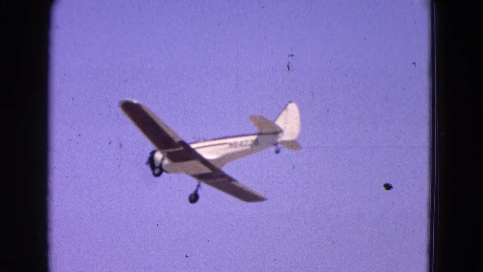 A small high-wing airplane is seen from below in 1964 8mm footage, angled across the frame with landing gear down.