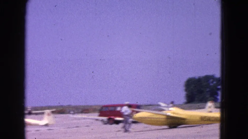 1964 Washington, DC home-movie frame showing small aircraft on a tarmac, including a yellow plane, with a red van and people in the background.