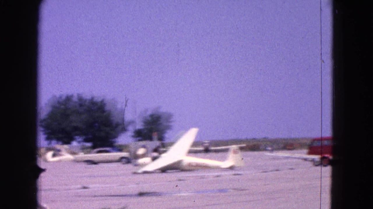 1964 Washington, DC airfield view with a white glider on the tarmac, parked cars, trees, and a red vehicle in the background.