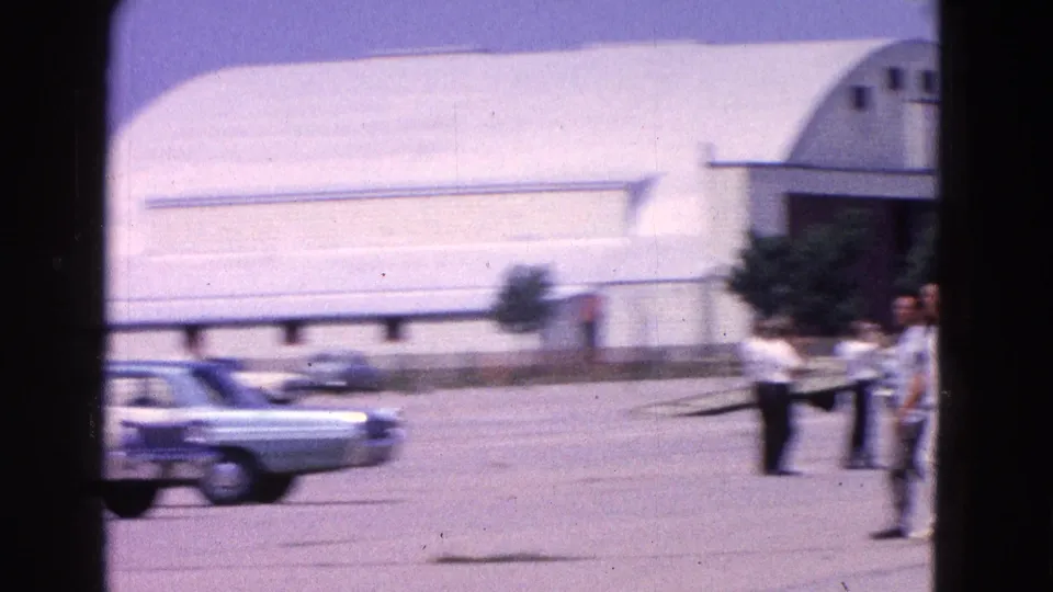 1964 Washington, DC airfield frame showing a large hangar, a parked car, part of a small aircraft, and people standing near the tarmac.
