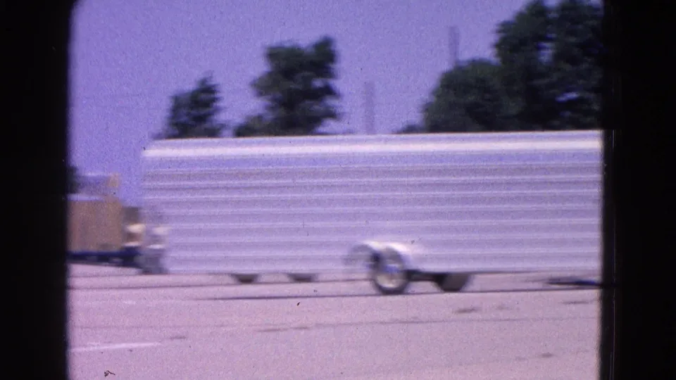 1964 Washington, DC home-movie frame showing a white trailer on the airfield tarmac with trees and low structures behind it.