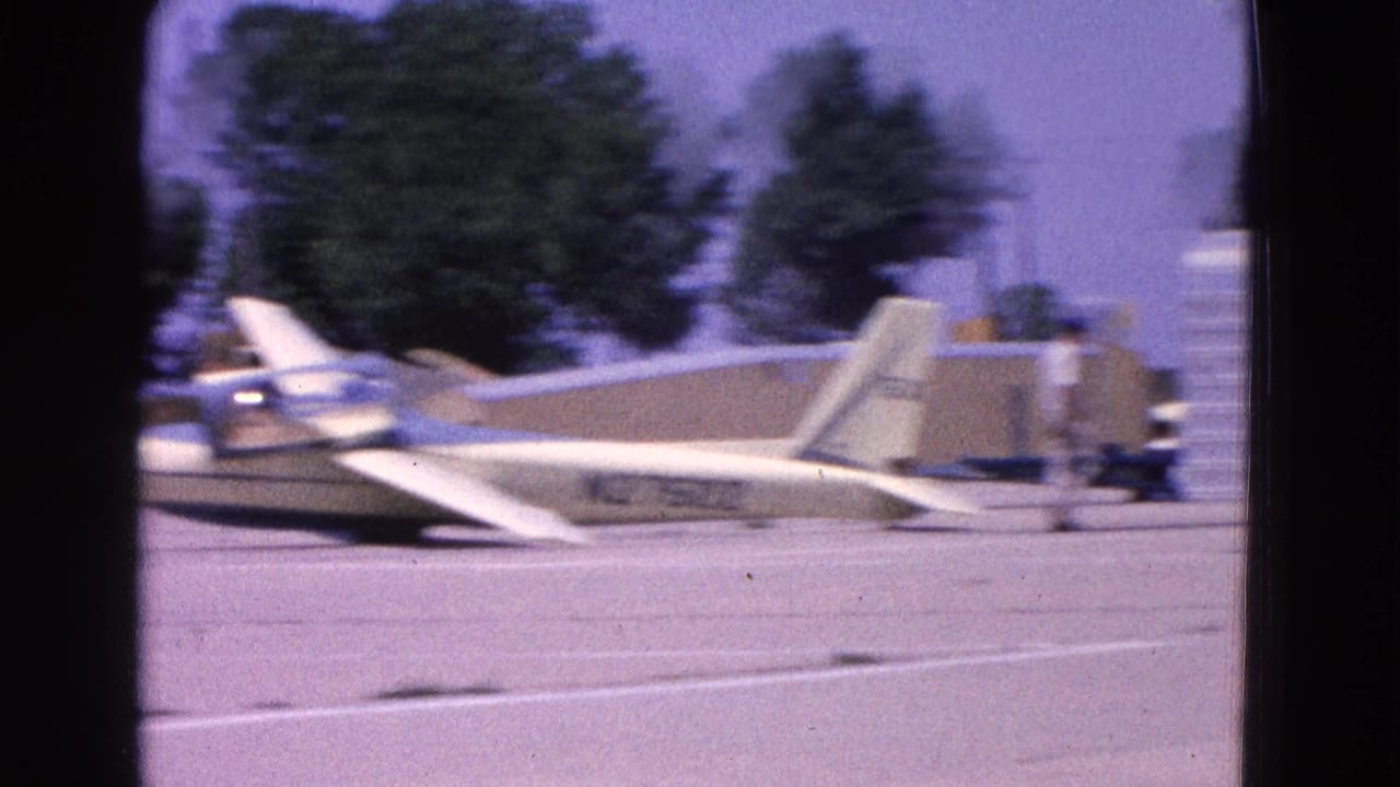 White glider on a runway with parked aircraft and trees beyond the tarmac.
