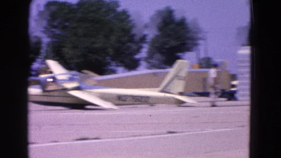 1964 Washington, DC 8mm frame showing a white glider on a sunlit runway with other small aircraft and trees in the background.