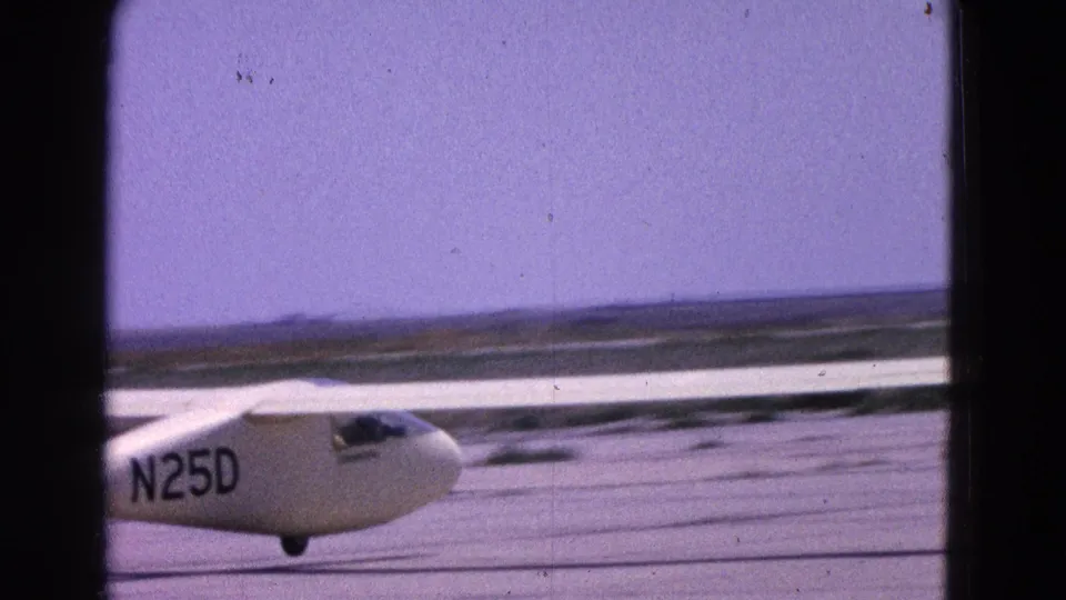1964 8mm home-movie frame of a white glider marked N25D moving along a runway, with long wings and a low horizon visible.