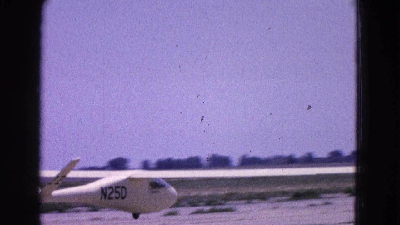 1964 Washington DC aviation footage showing the white glider N25D on a runway, framed through visible 8mm gate edges.
