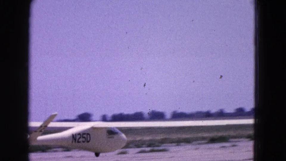 1964 Washington DC aviation footage showing the white glider N25D on a runway, framed through visible 8mm gate edges.