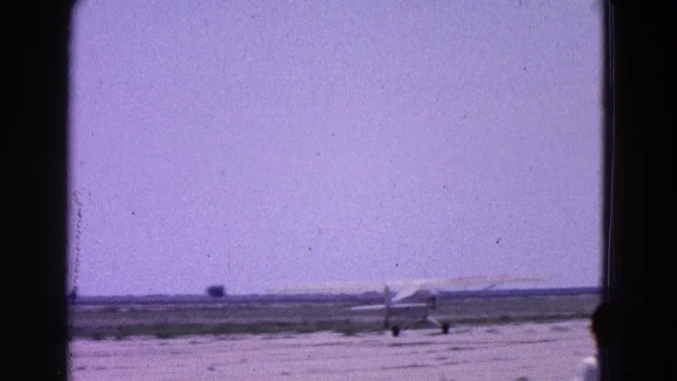A distant white glider is seen on the runway while a person appears partly at the right edge of the 1964 home-movie frame.