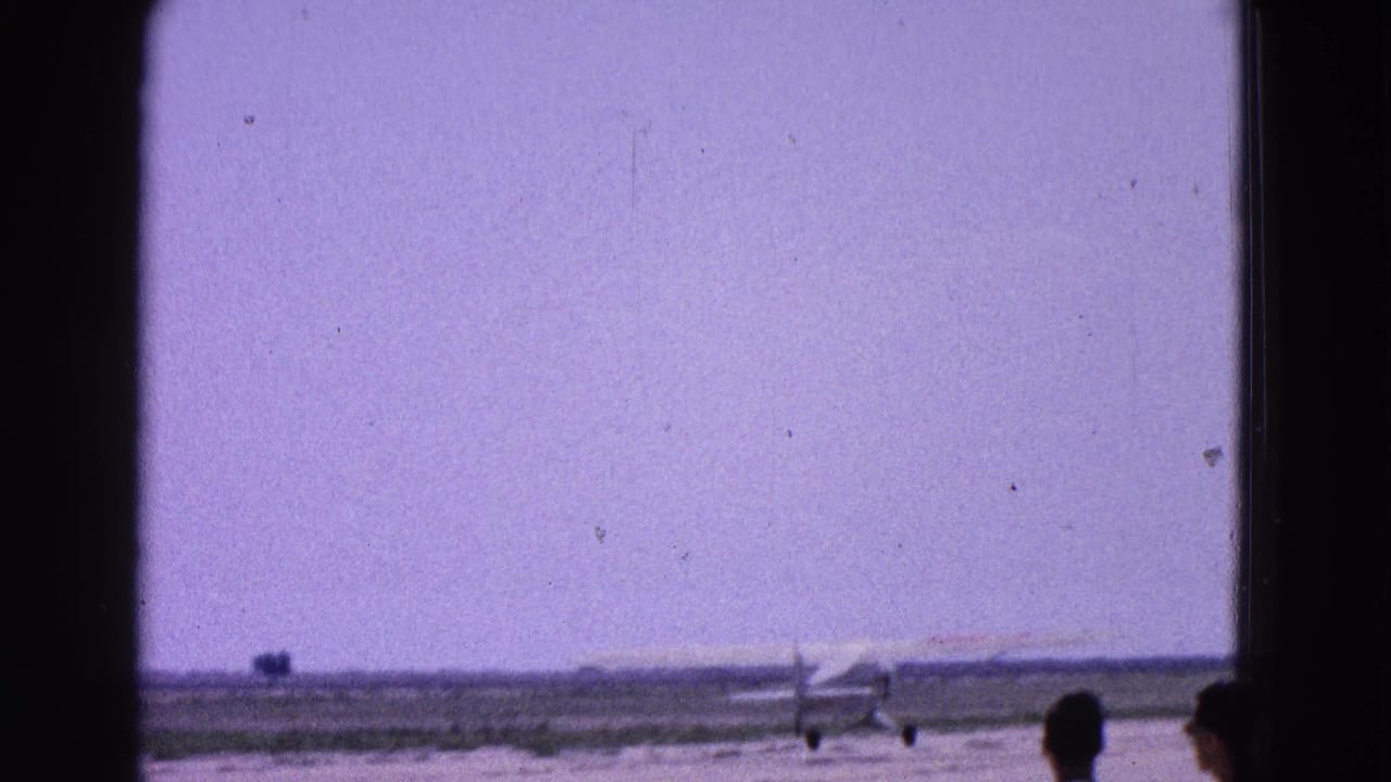 1964 Washington, DC home-movie frame showing a small propeller plane on a runway beneath a hazy purple sky.