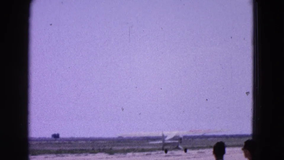 1964 Washington, DC home-movie frame showing a small propeller plane on a runway beneath a hazy purple sky.