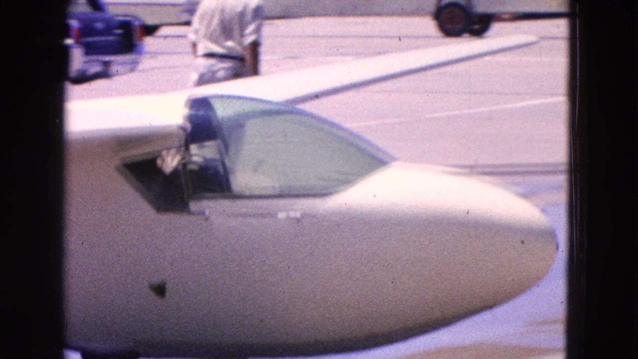 1964 Washington DC home-movie frame showing a small white plane nose, cockpit canopy, tarmac, and parked cars behind it.