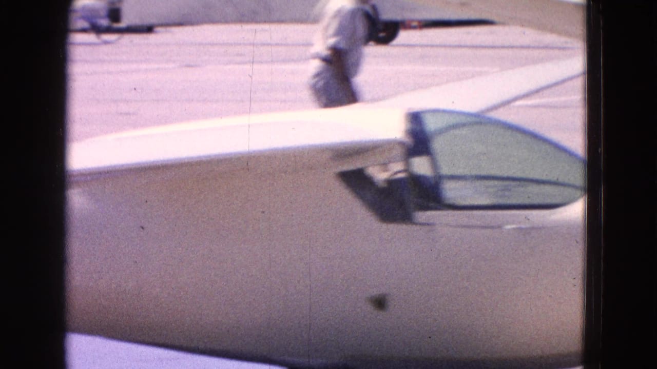 1964 Washington DC airport frame with the small aircraft cockpit, white fuselage, tarmac, and a walking adult in the background.