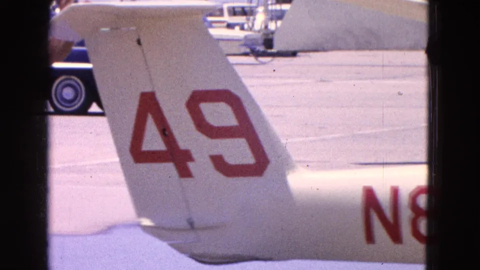 1964 Washington DC airport frame showing the small plane tail marked 49, partial registration, tarmac, and parked cars.