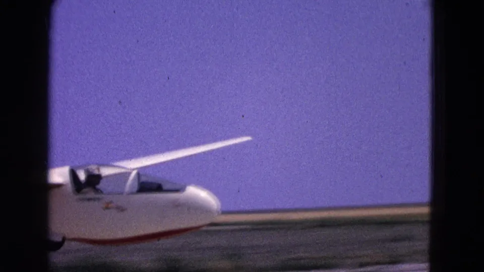 Close 8mm frame of the glider passing on the runway, showing the nose, cockpit, wing root, and mid-century home-movie color shift.