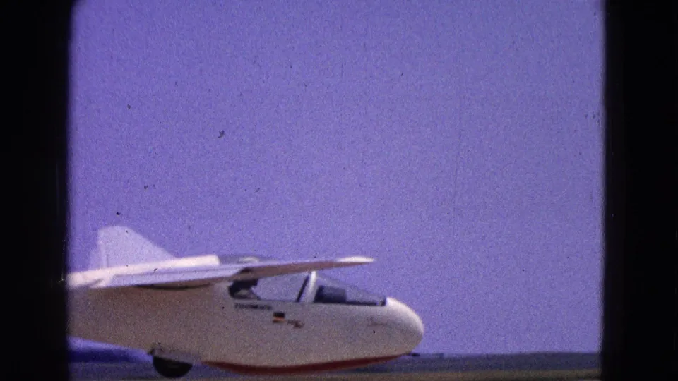 The white glider appears farther along the runway in the 1964 home-movie footage, with wing, tail, cockpit area, and blue sky visible.