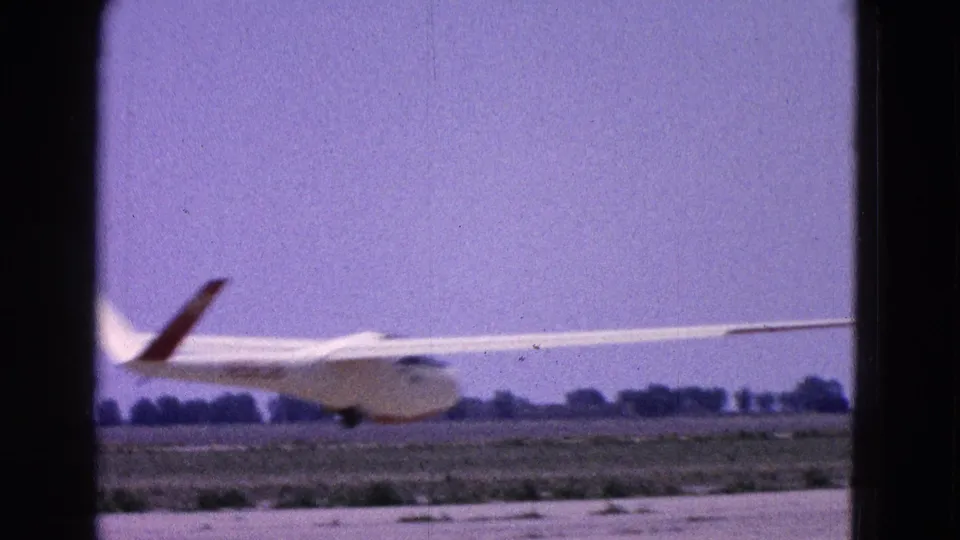A low, soft 8mm view shows the glider near liftoff with runway, grass, and distant trees behind it.