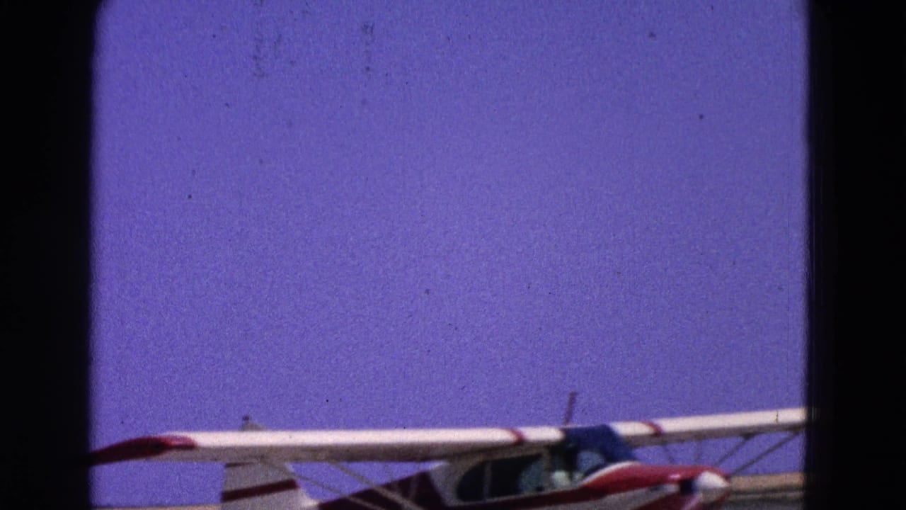 1964 Washington, DC 8mm frame showing a small white-and-red aircraft low in the frame near a runway under a clear blue sky.