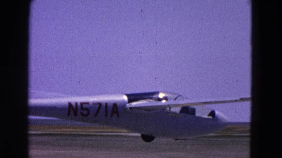 1964 Washington, DC 8mm frame showing the rear of a white small aircraft marked N571A as it passes across a blue-sky runway view.