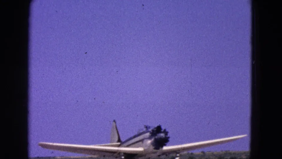 1964 Washington, DC home-movie frame showing a white single-engine propeller airplane with blue striping near the runway edge.